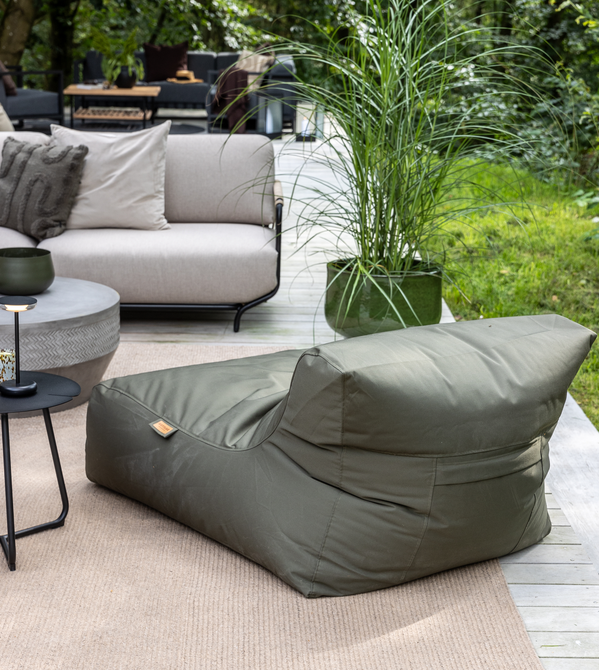 Grøn sækkestol/beanbag på en træterrasse ved siden af en beige sofa, lille sort sidebord med lys og høj prydgræs i krukke; frodig have i dagslys.