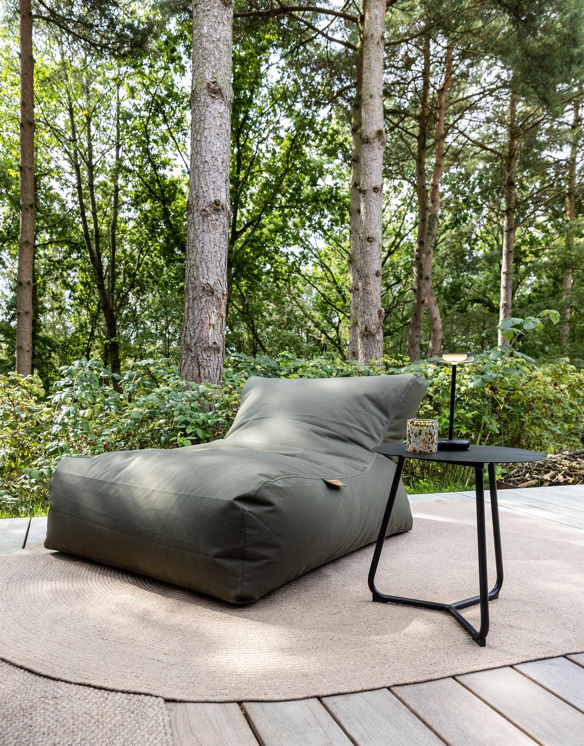 Grøn sækkestol på træterrasse med rund beige tæppe og lille sort sidebord med lampe og kop, omgivet af skov i dagslys.