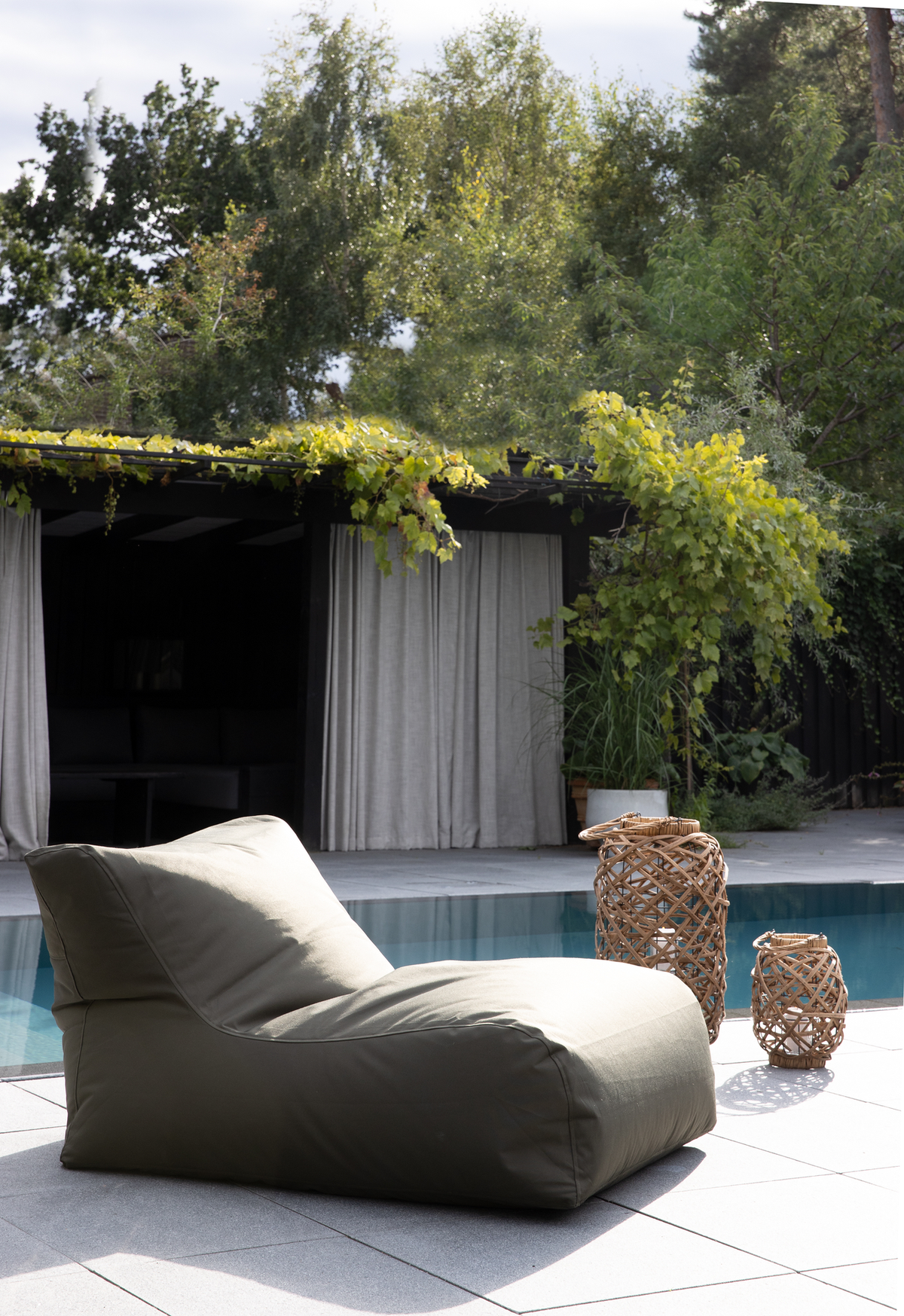 Grøn loungestol ved en pool på terrasse med to flettede lanterner og pergola med grønne ranker og gardiner i baggrunden.