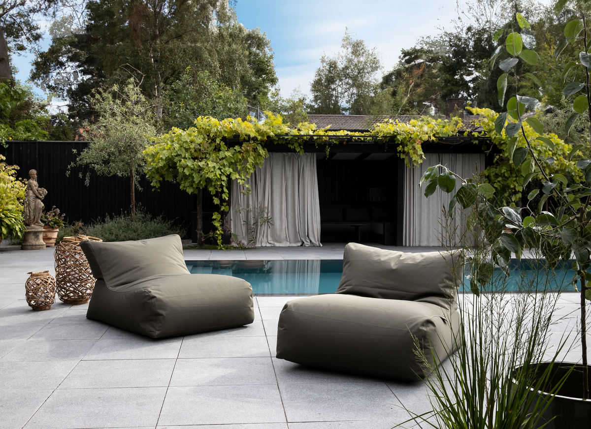 Terrasse med to olivengrønne loungestole ved en pool, flettede lanterner og pergola med lyse gardiner i dagslys.