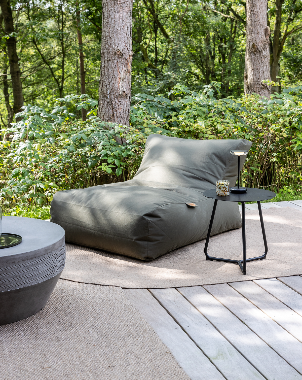 Grøn sækkestol‑lounge på træterrasse med rund måtte, sort sidebord med bærbar lampe og lille lyslygte, skov i baggrunden.