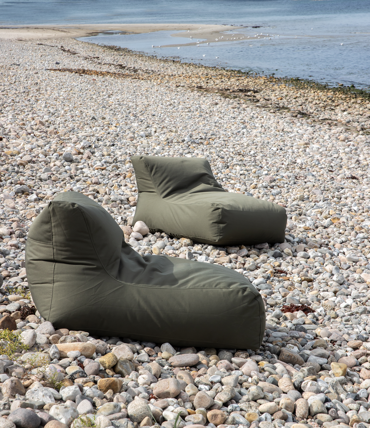 To olivengrønne sækkestols-loungestole står på en stenstrand ved havet.