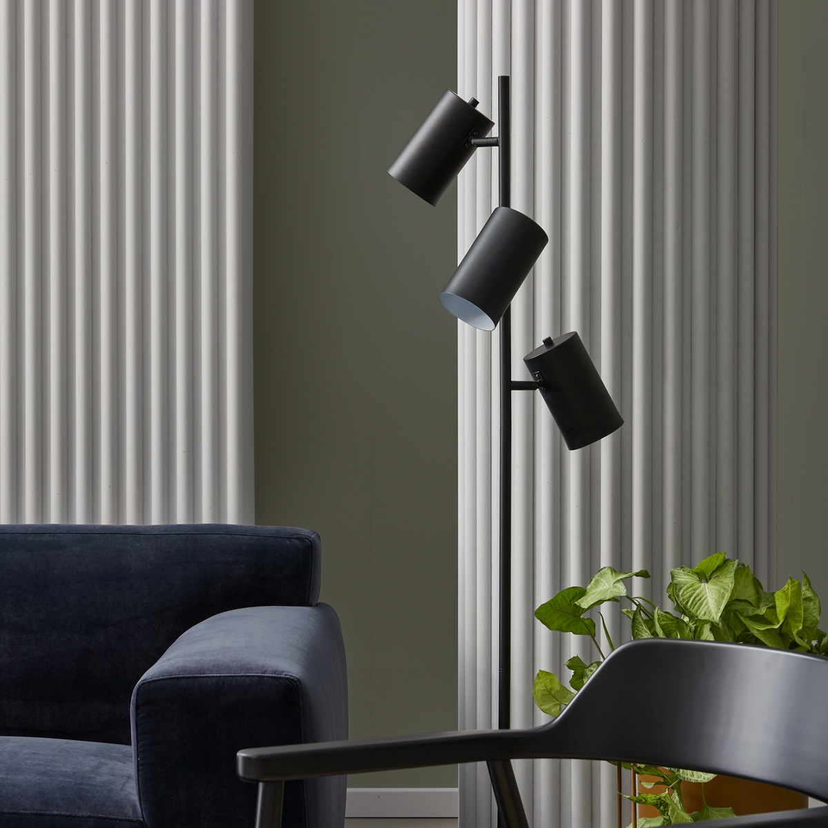 Moderne stue med sort gulvlampe med tre spotlights, mørkeblå sofa, sort stol og grøn potteplante foran riflede lysegrå paneler.
