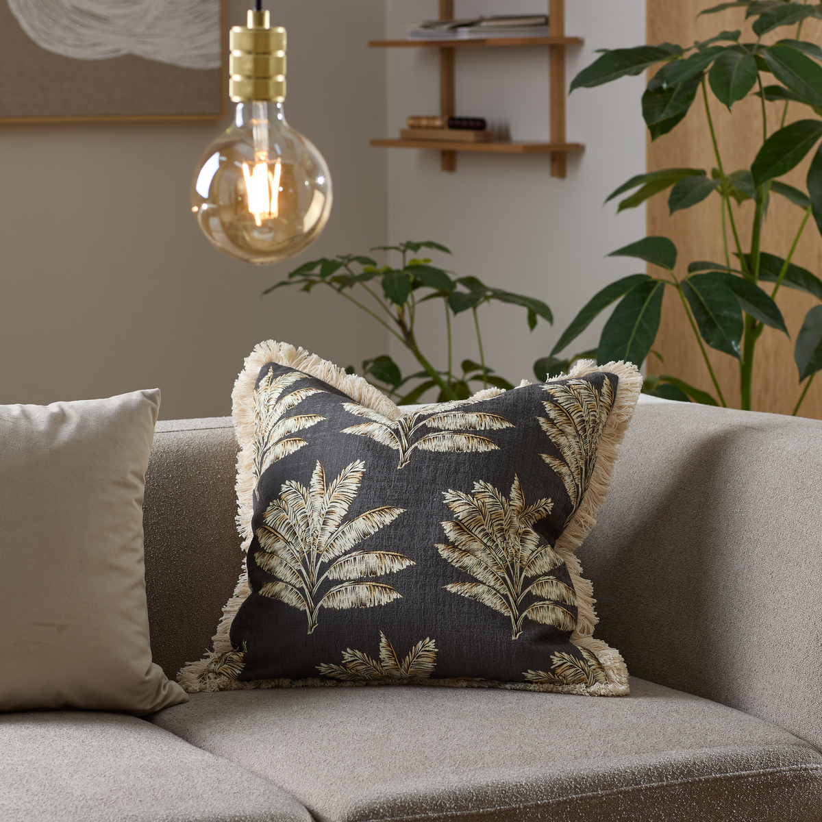 Beige sofa med pyntepude med palmeblade i sort og beige, tændt pære og grønne planter i en moderne stue.