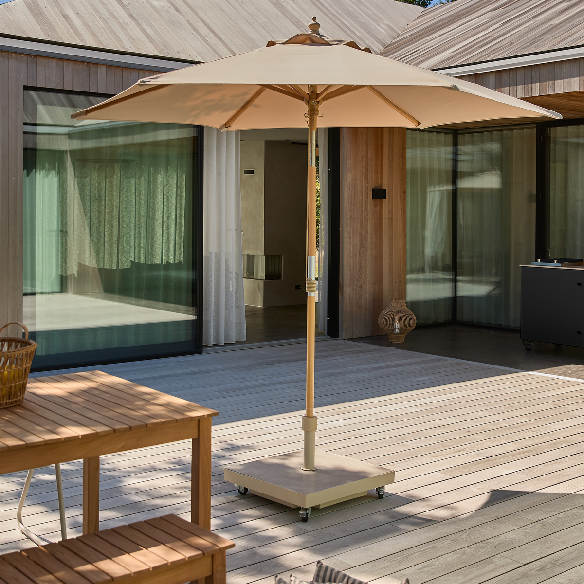 Beige parasol med base på hjul på en træterrasse foran et moderne hus med store skydedøre og gardiner; træbord i forgrunden.