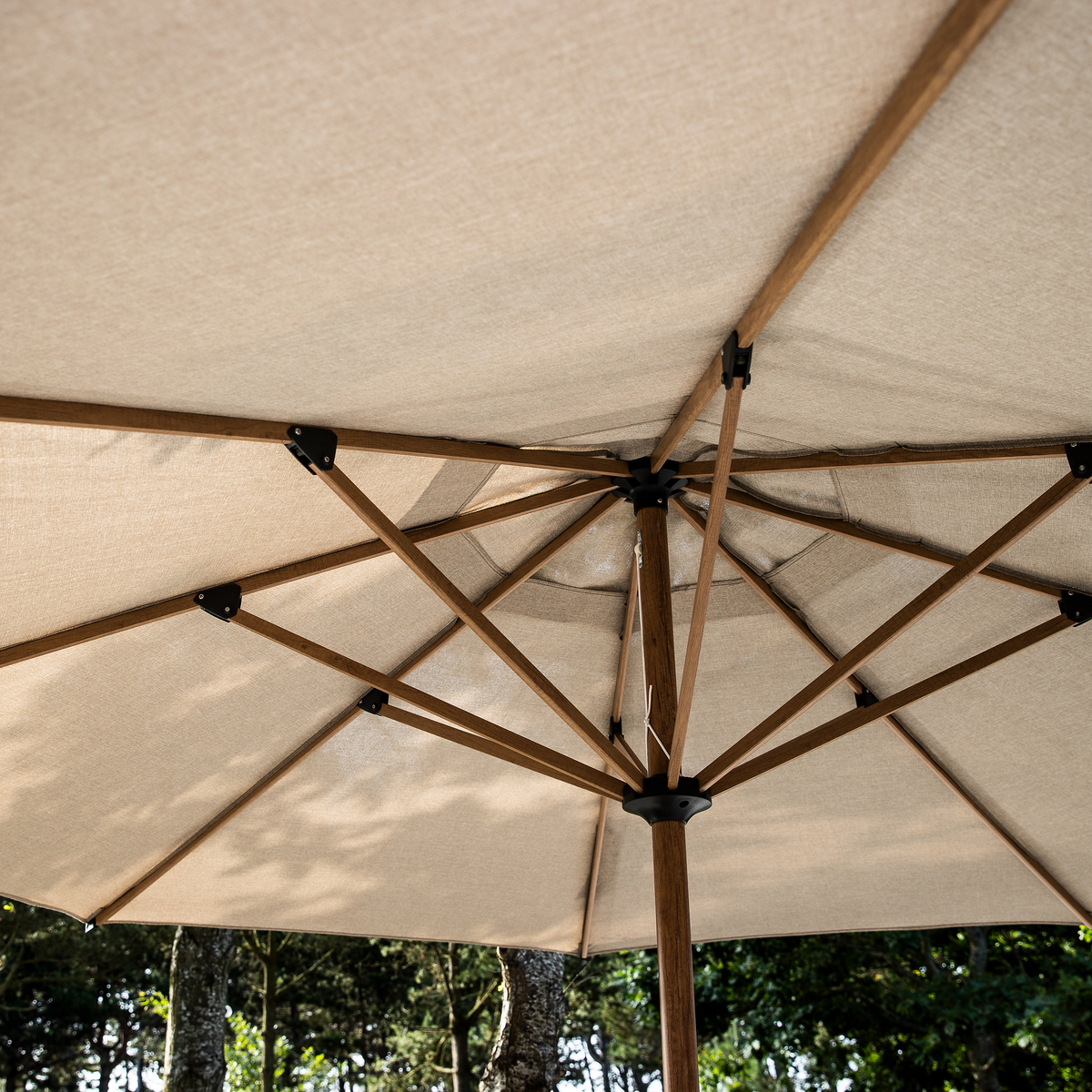 Undersiden af en beige udendørs parasol med træstang og eger i dagslys blandt træer.