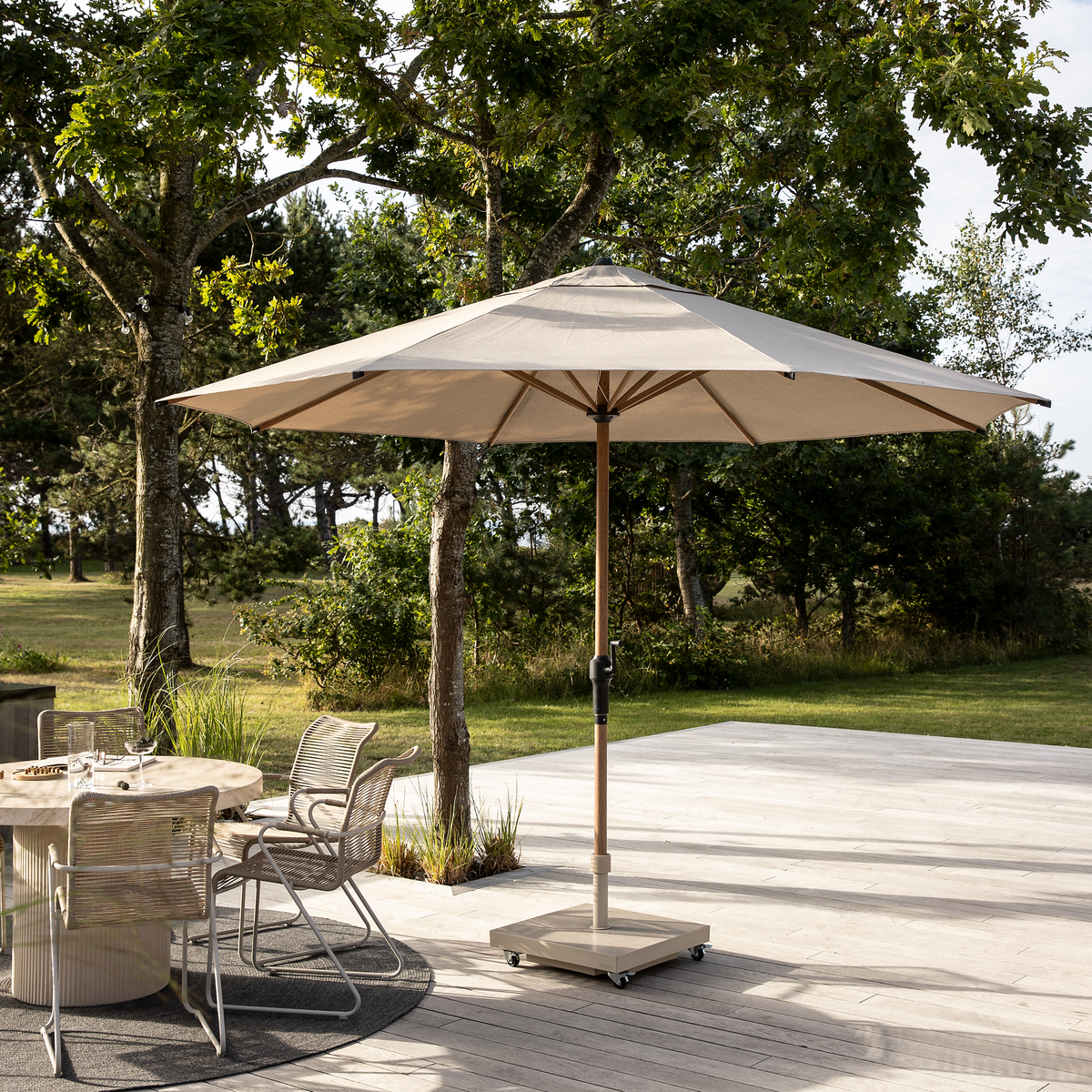 Terrasse med beige parasol på hjulbase, rundt spisebord og stole i havemiljø i dagslys.