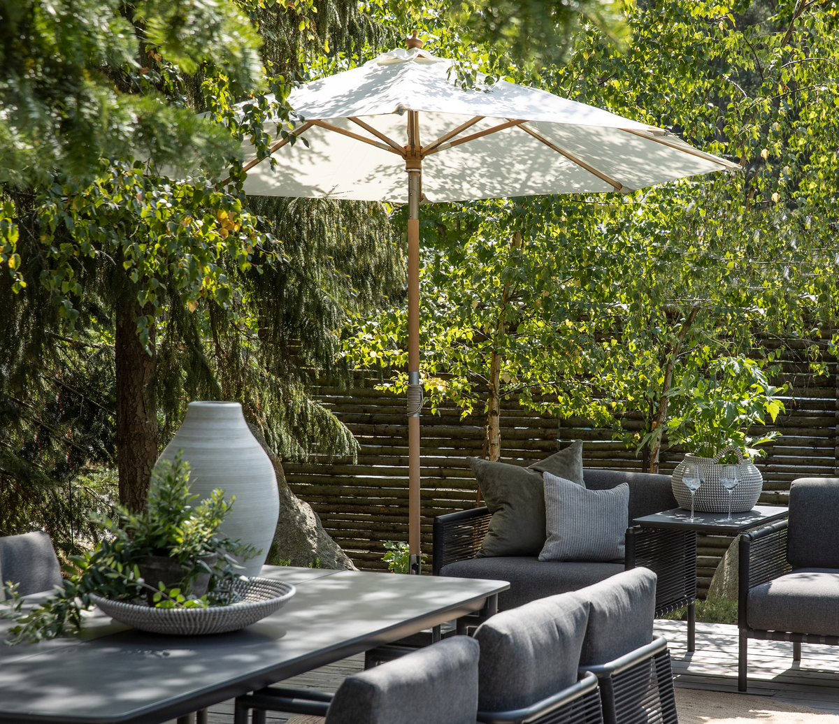 Sommerlig terrasse med hvid parasol, grå loungemøbler med puder, grønne planter og en vase på bordet, omgivet af træer og træhegn i dagslys.