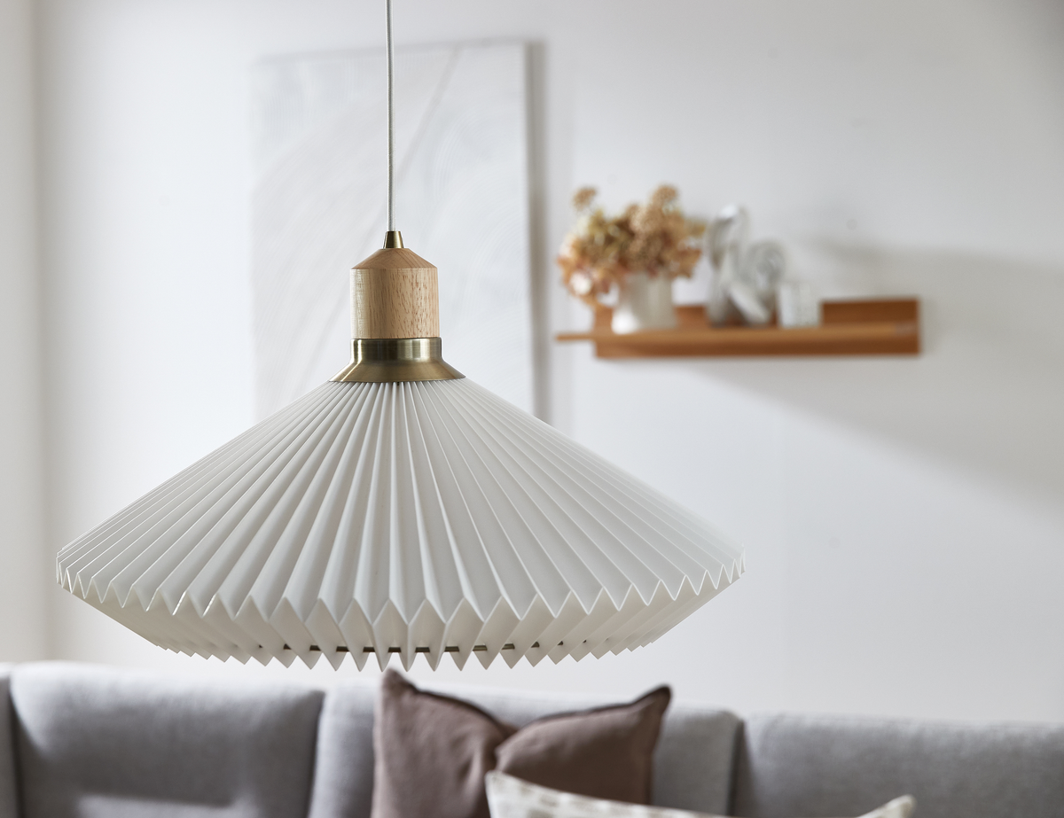 Hvid plisseret pendellampe med træ- og messingdetalje over grå sofa; hylde med vase og dekorationer i baggrunden.