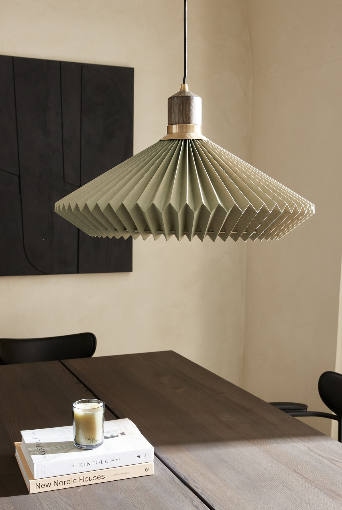 Plisseret grøn pendellampe over mørkt spisebord med bøgerne New Nordic Houses og The Kinfolk Home samt et duftlys med teksten Lemon Verbena; sort vægkunst i baggrunden.