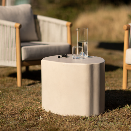 Lille beige udendørs sidebord med karaffel og drikkeglas samt et par brombær, mellem to loungestole på tør græs i varmt dagslys.