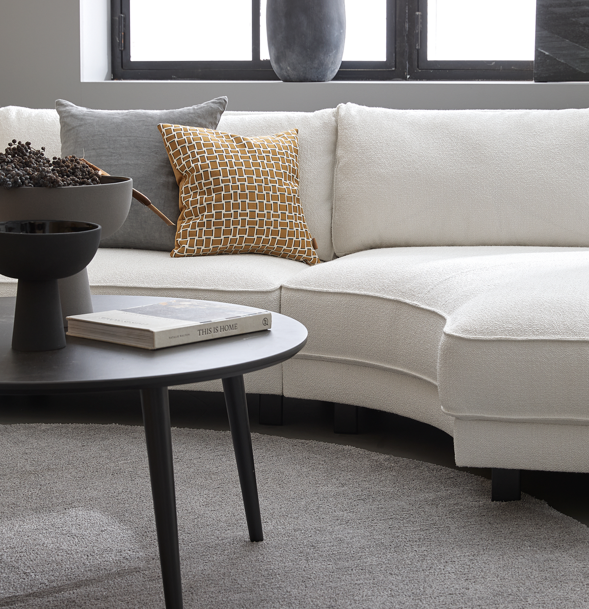 Sort sofabord med skål og bogen 'This Is Home' foran en lys, buet sofa med grå og sennepsgul pude i en moderne stue.