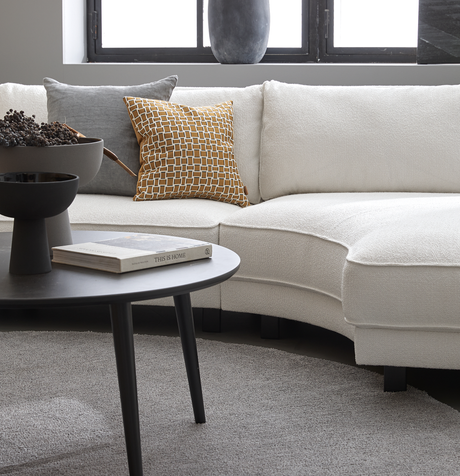 Sort sofabord med skål og bogen 'This Is Home' foran en lys, buet sofa med grå og sennepsgul pude i en moderne stue.