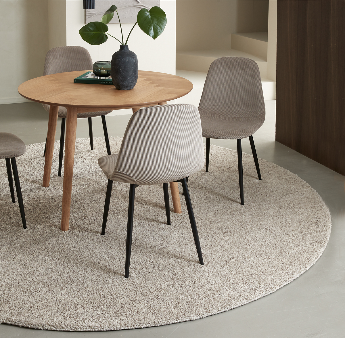 Rundt egetræsbord med fire beige polstrede stole på et lyst rundt tæppe; sort vase med grønne blade og et lille lys på bordet i en minimalistisk spisestue.