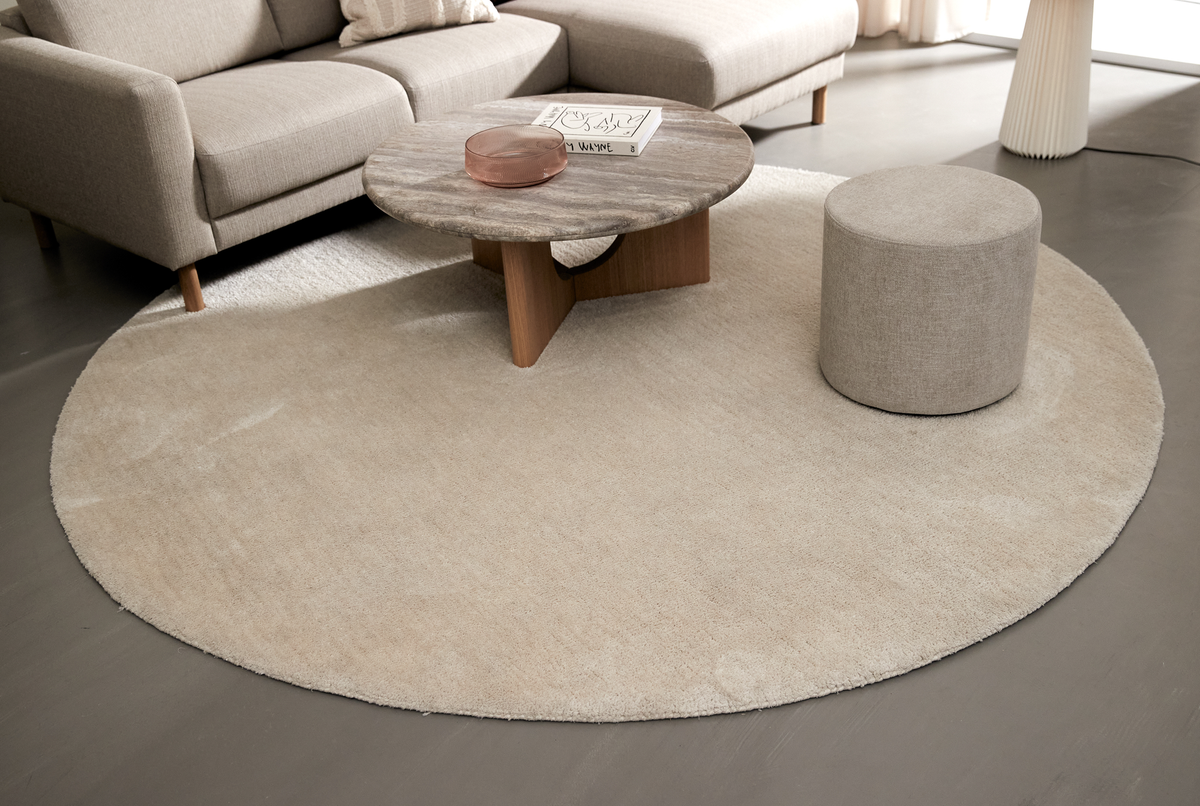 Beige rundt tæppe i en moderne stue med sofa og rundt sofabord i sten; på bordet ligger bogen ”DRAWING” og en rosa glasskål.