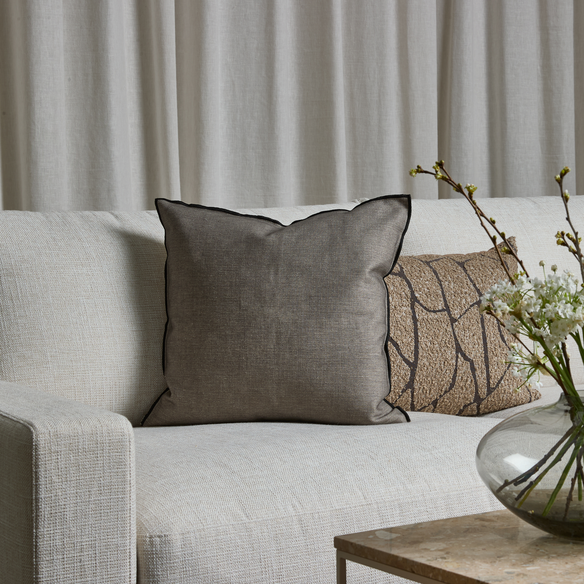 Lys sofa med grå pude med sort kant, beige mønstret pude og glasvase med hvide blomster på stenbord foran lyse gardiner.