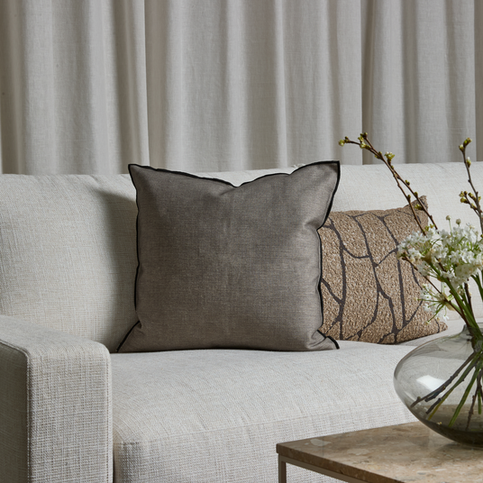 Lys sofa med grå pude med sort kant, beige mønstret pude og glasvase med hvide blomster på stenbord foran lyse gardiner.