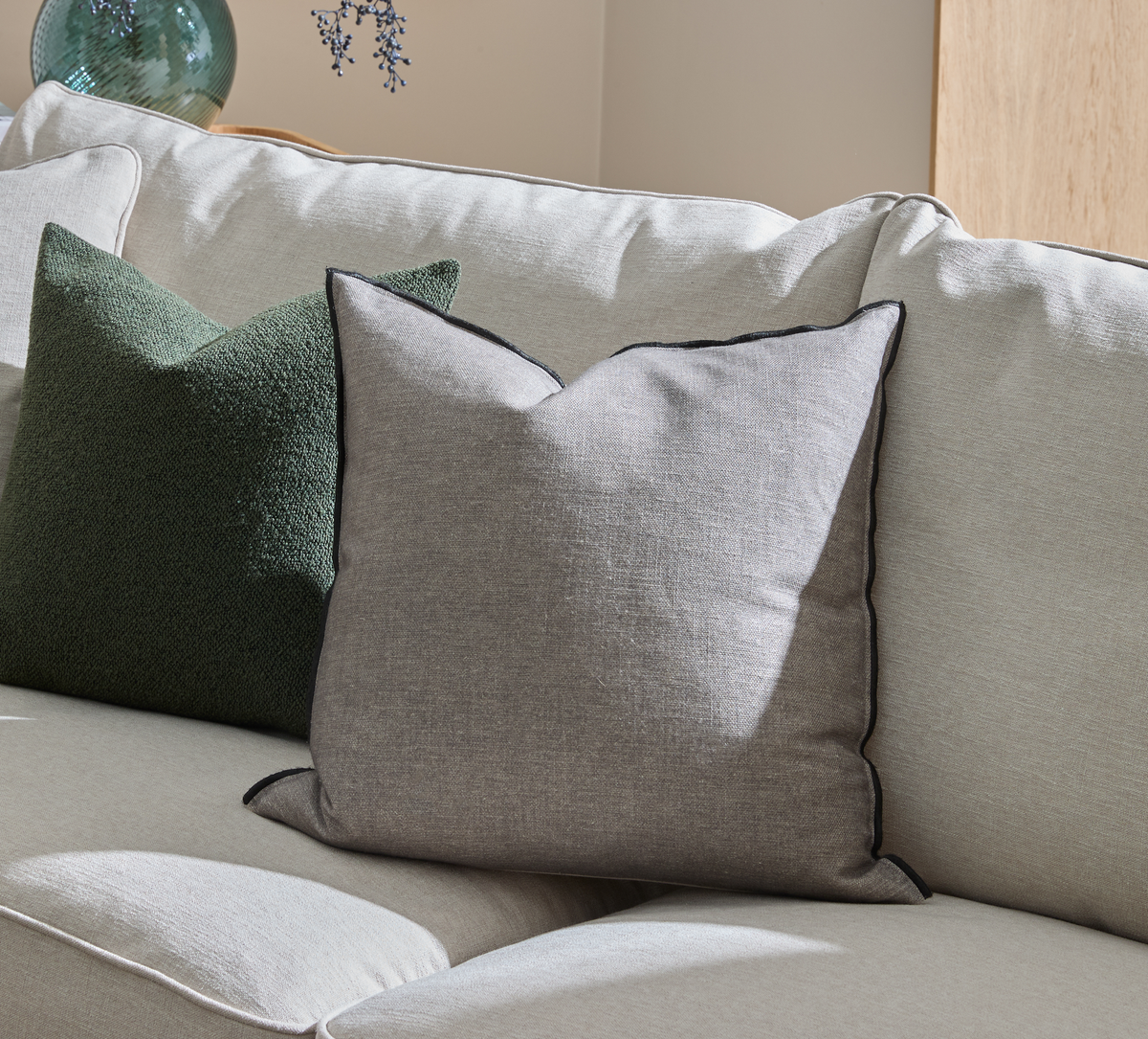 Grå pyntepude med mørk piping på en lys beige sofa ved siden af en grøn pude i et solbelyst stuehjørne.