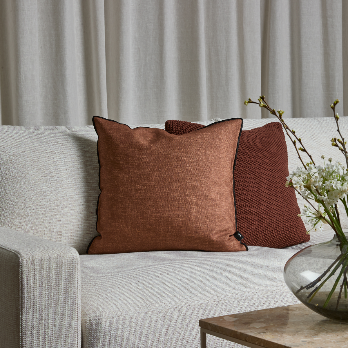 Beige sofa med to rustfarvede puder, den forreste med sort kantsyning, hvide gardiner bagved og en glasvase med hvide blomster på et stenbord.