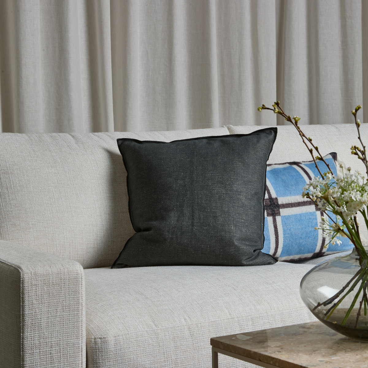 Lys sofa med mørkegrå pude, blåternet pude og glasvase med hvide grene på et sidebord foran lyse gardiner.