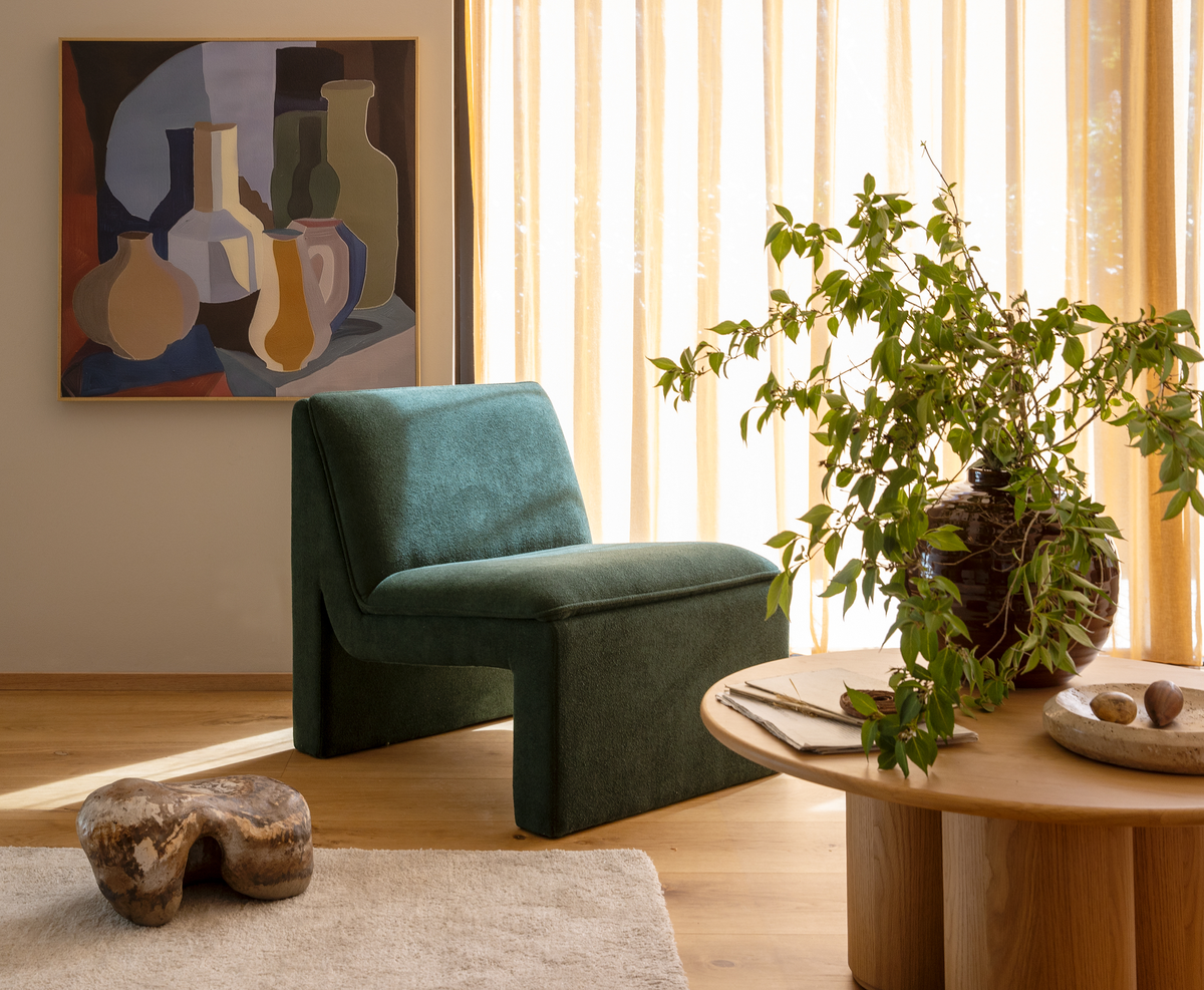 Lyst stueinteriør med mørkegrøn loungestol, rundt sofabord i træ med grøn plante i vase, abstrakt maleri på væggen og lyse, transparente gardiner i dagslys.