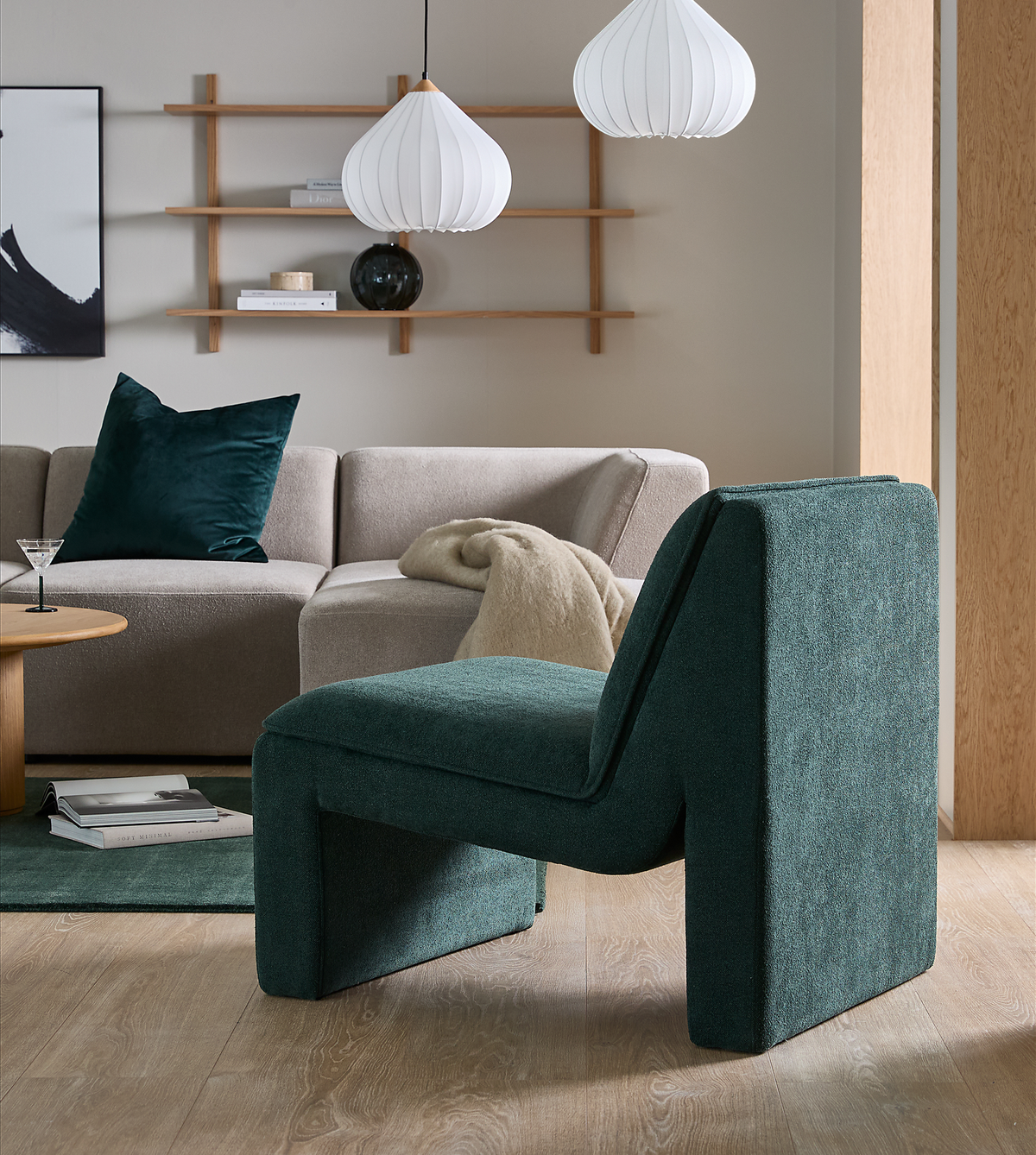 Moderne stue med skulpturel mørkegrøn loungestol i fokus, beige modulsofa, hvide pendler, rundt træbord og væghylder – rolig, neutral palet med grøn måtte og plaid.