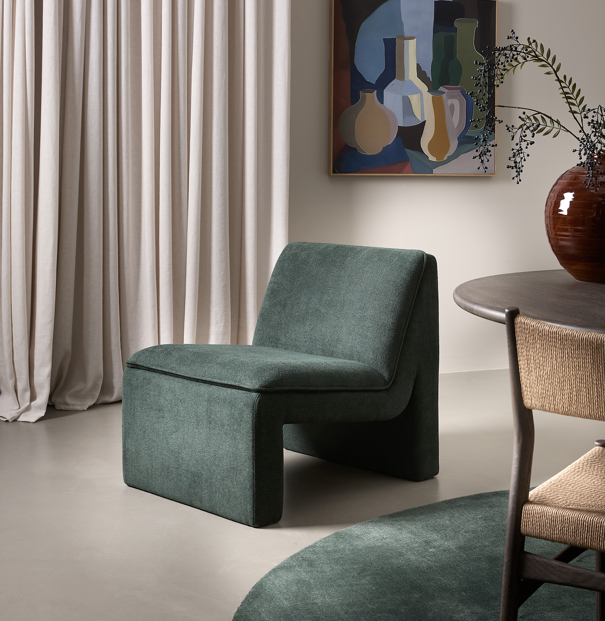 Moderne grøn loungestol i et neutralt rum med beige gardiner, abstrakt stilleben-vægkunst, rund grøn tæppe og kant af spisebord med fletstol i blødt dagslys.