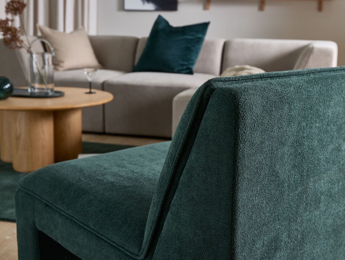 Nærbillede af en mørkegrøn polstret lænestol foran en beige modulsofa med puder og et rundt sofabord i træ med glasvase og cocktailglas i en lys stue.