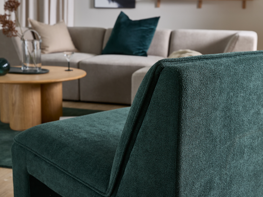 Nærbillede af en mørkegrøn polstret lænestol foran en beige modulsofa med puder og et rundt sofabord i træ med glasvase og cocktailglas i en lys stue.