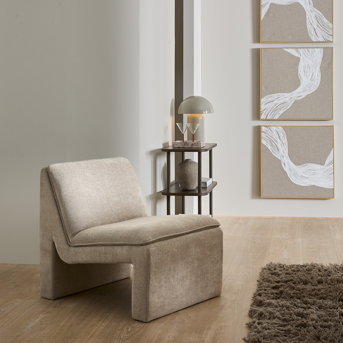Beige loungestol i en minimalistisk stue med sidebord, kuppelformet bordlampe og tre abstrakte billeder på væggen samt et brunt ryetæppe på trægulv.