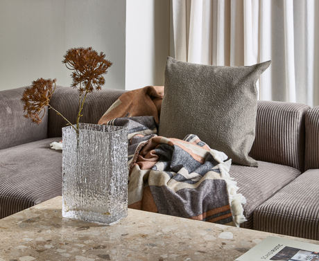 Hjørne af en gråriflet sofa med beige pude og mønstret plaid; glasvase med tørrede grene på et terrazzobord og et magasin med titlen Scandi Rustic i hjørnet.