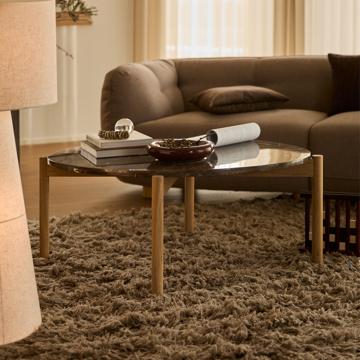 Moderne stue med rundt marmorsofabord med træben på en langhåret tæppe, neutral sofa, gulvlampe og bøger, blødt dagslys gennem gardiner.