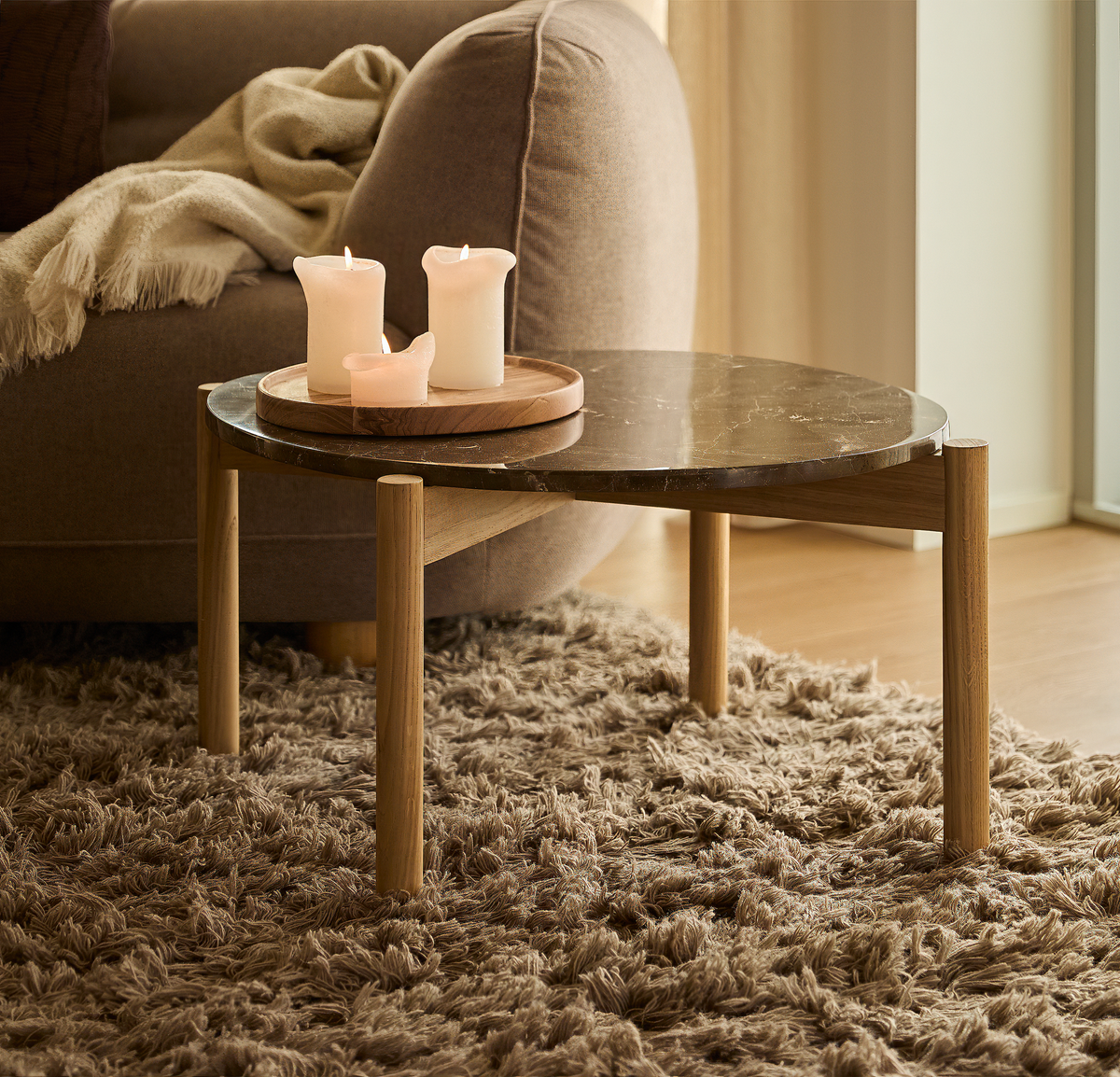 Lavt sofabord med mørk marmorlignende bordplade og træben på brun højluvet tæppe, med tændte lys på en rund træbakke ved en beige sofa i dagslys.