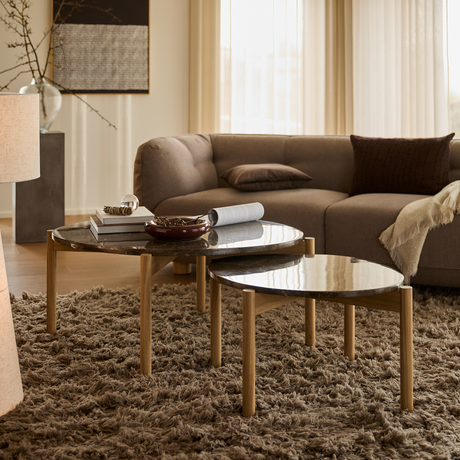 Stue med to runde sofaborde i mørk sten og egetræ på brun ryatæppe foran beige modulsofa med puder og plaid, bløde gardiner og varm belysning.