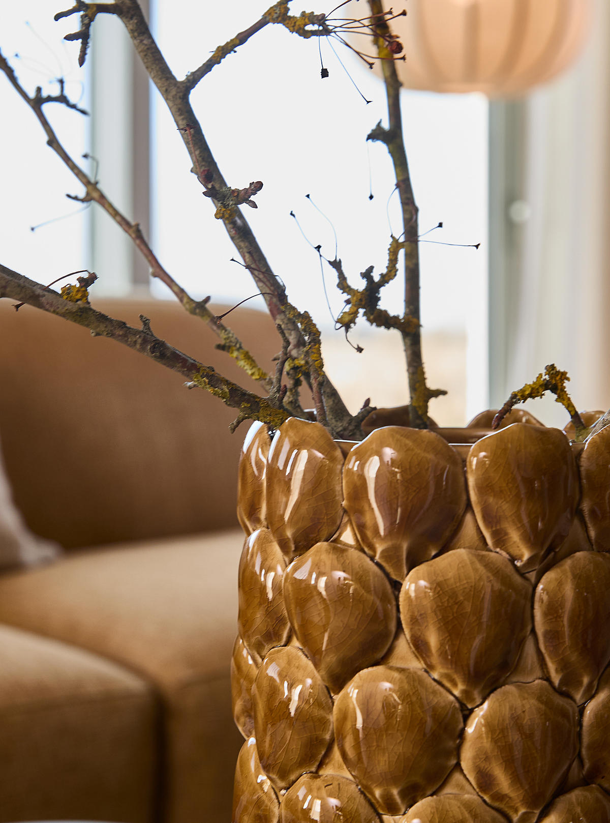 Nærbillede af brun glaseret keramikvase med bladmønster, fyldt med tørre grene, i en lys stue med beige sofa.