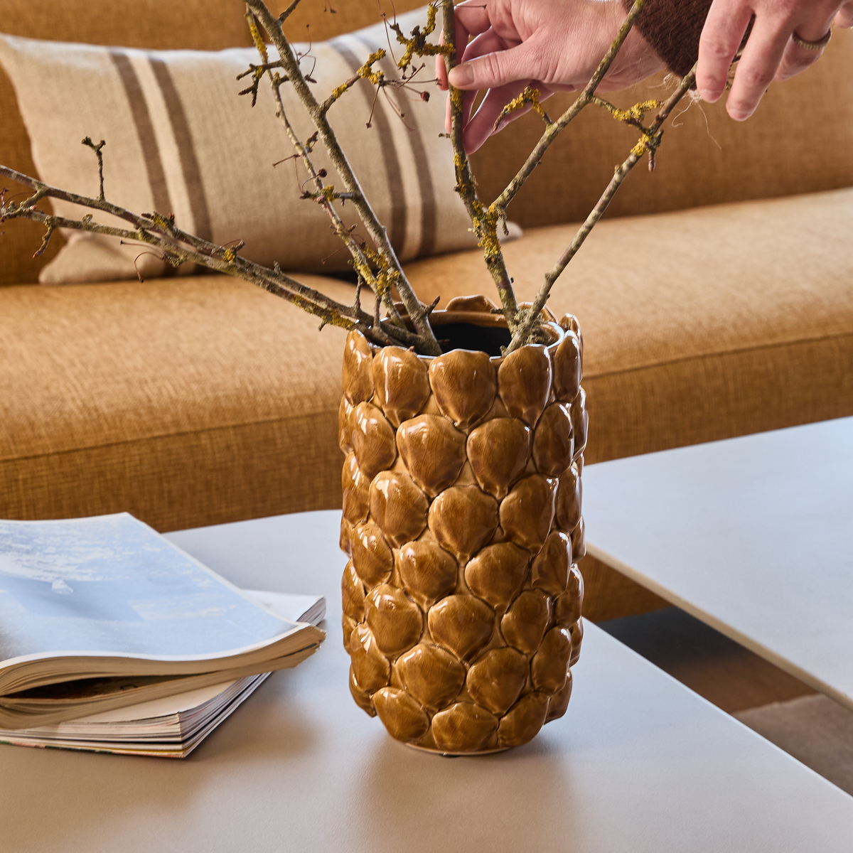 Brun keramisk vase med tekstureret mønster på sofabord; en hånd arrangerer tørre grene, med sofa og pude i baggrunden.