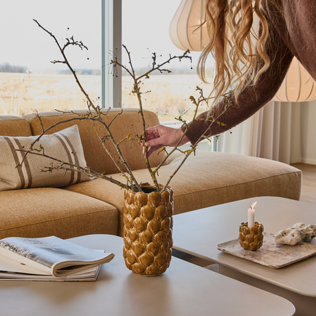 Hånd der arrangerer grene i en brun, struktureret vase på sofabord; beige sofa, tændt lys og åben bog i et lyst stuemiljø.