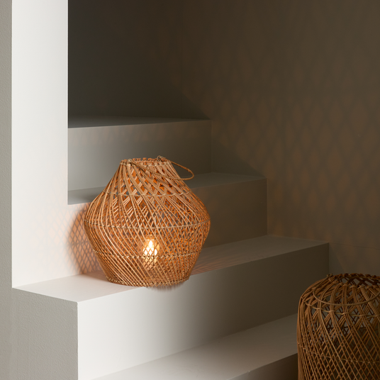 Flettet rattanlampe med varmt lys på hvide trappetrin, der kaster diamantmønstrede skygger på væggen.
