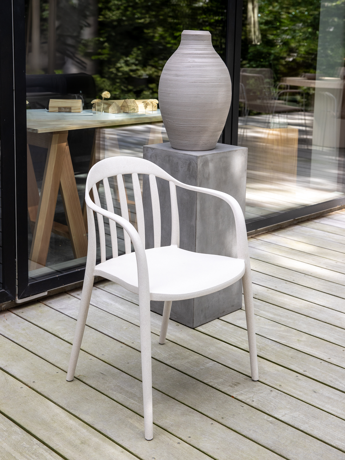 Hvid udendørs stol på træterrasse ved siden af en stor grå keramisk vase på betonsokkel, med glasfacade og grøn beplantning i baggrunden.