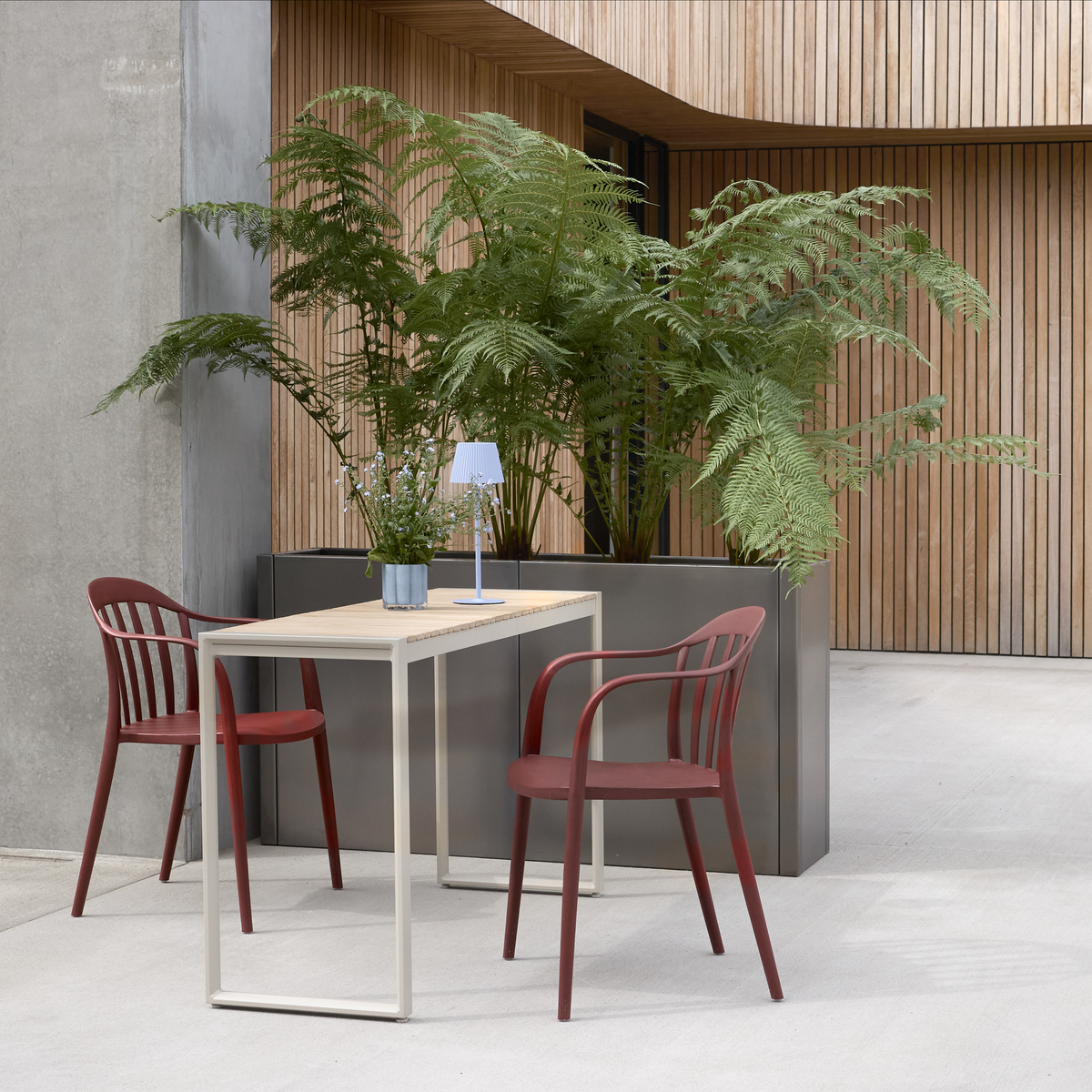 Moderne udendørs terrasse med to røde stole, lyst rektangulært bord, lille bærbar bordlampe og potteplante foran træbeklædt facade og store bregner i plantekasse.