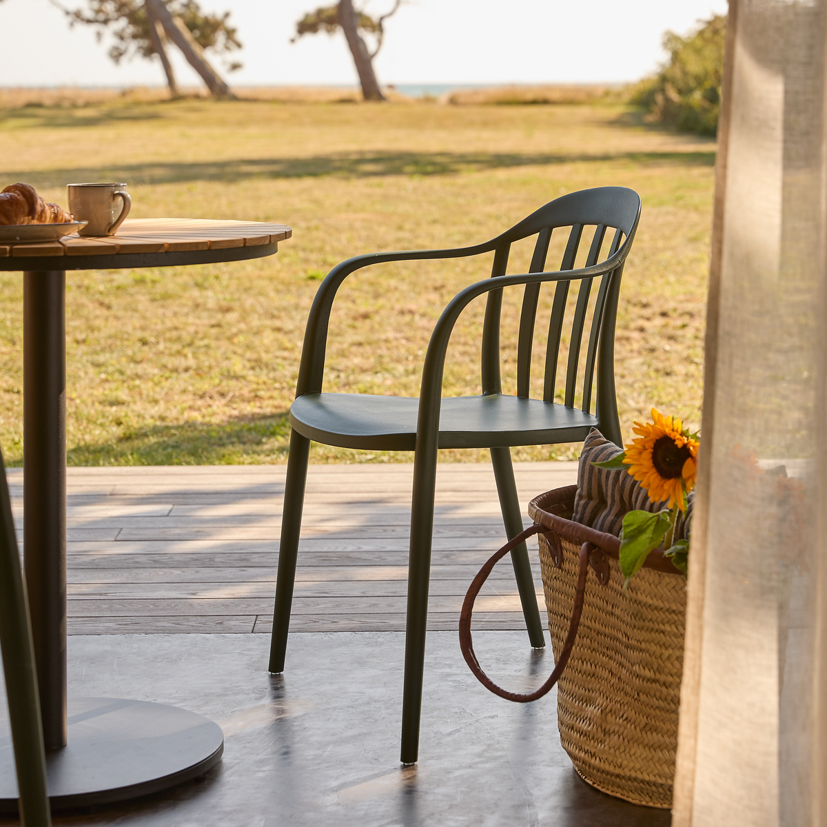 Grøn udestol ved et rundt terrassebord med kaffekop og croissant; ved siden af står en fletkurv med solsikke, solrig græsplæne og hav i baggrunden.