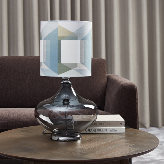 Røget glasbordlampe med geometrisk mønstret skærm på sofabord ved siden af bøgerne The Kinfolk Table og A Frame for Life, foran brun sofa og beige gardiner.
