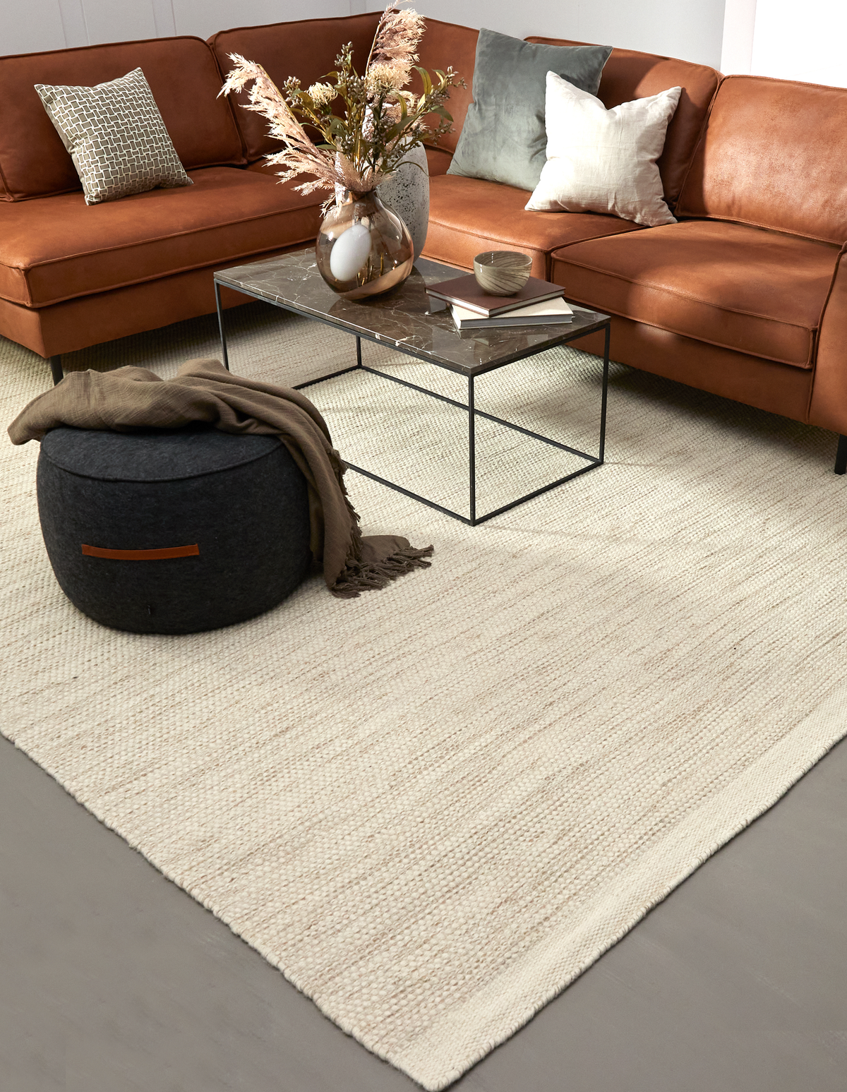 Lys beige vævet tæppe i en stue med cognacfarvet hjørnesofa, marmorbord med sort metalstel, vase med tørrede grene og en mørk puff med plaid.