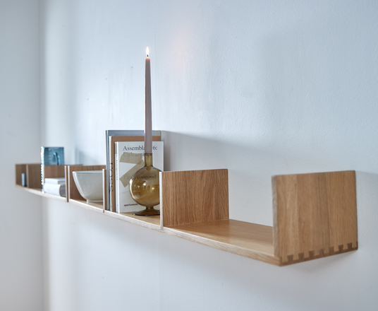 Vægmonteret hylde i lyst træ med rum, dekoreret med glas, skål, bøger (en med titlen “Assemblage 6:”) og en tænd lysestage.