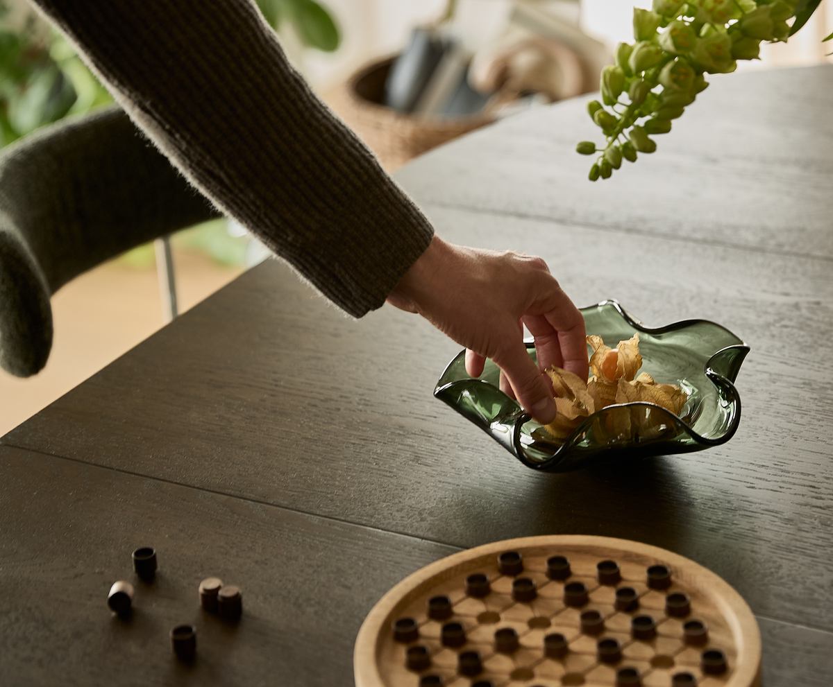 Hånd der tager snacks fra grøn glasskål på mørkt træbord ved siden af rundt brætspil; hjemmemiljø med planter i dagslys.
