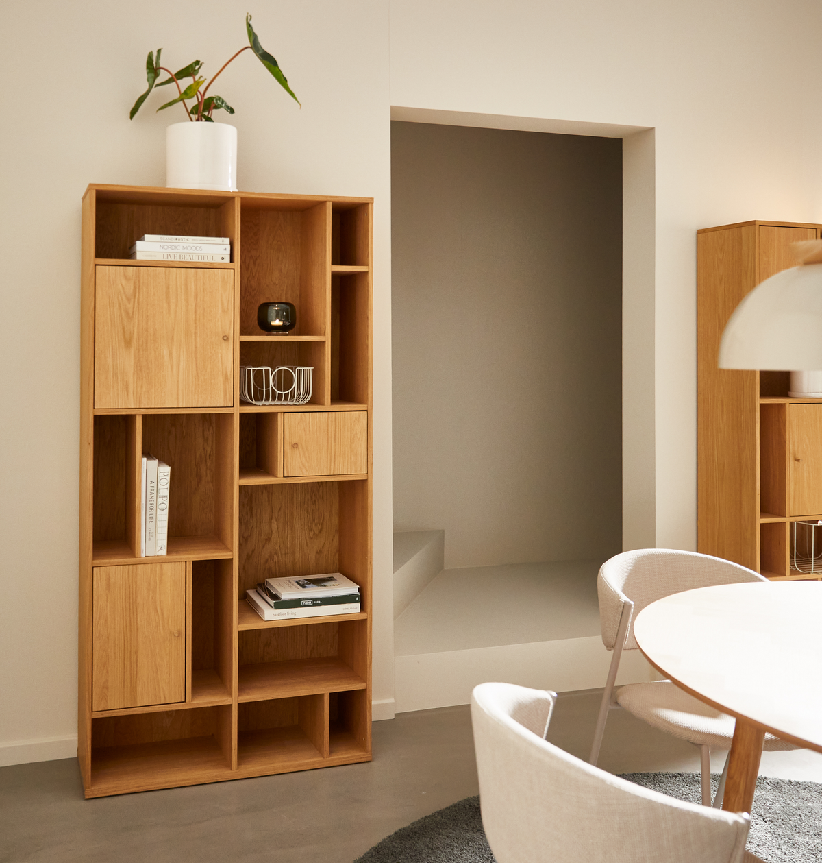 Lyst skandinavisk spiseområde med modulopbygget reol i eg; på hylden ses bøger med titlerne “NORDIC MOODS” og “NORM ARCHITECTS”, en hvid potte med grøn plante og en hvid trådkurv; rundt bord med polstrede stole og hvid pendellampe i neutrale nuancer.