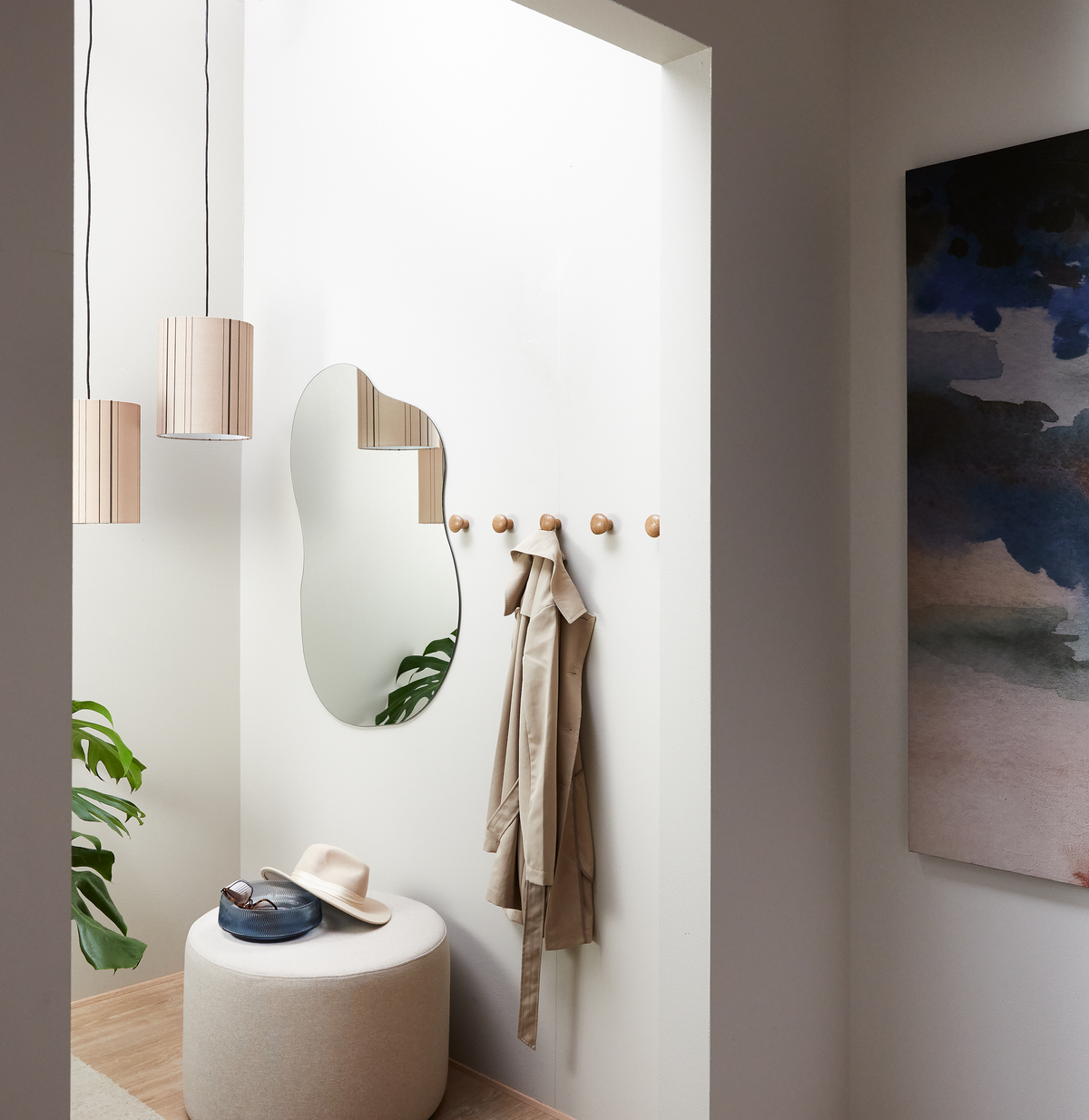 Lys entré med organisk spejl, træknager med beige trenchcoat, rund puf med hat og solbriller, hængende lamper og grønne planter.