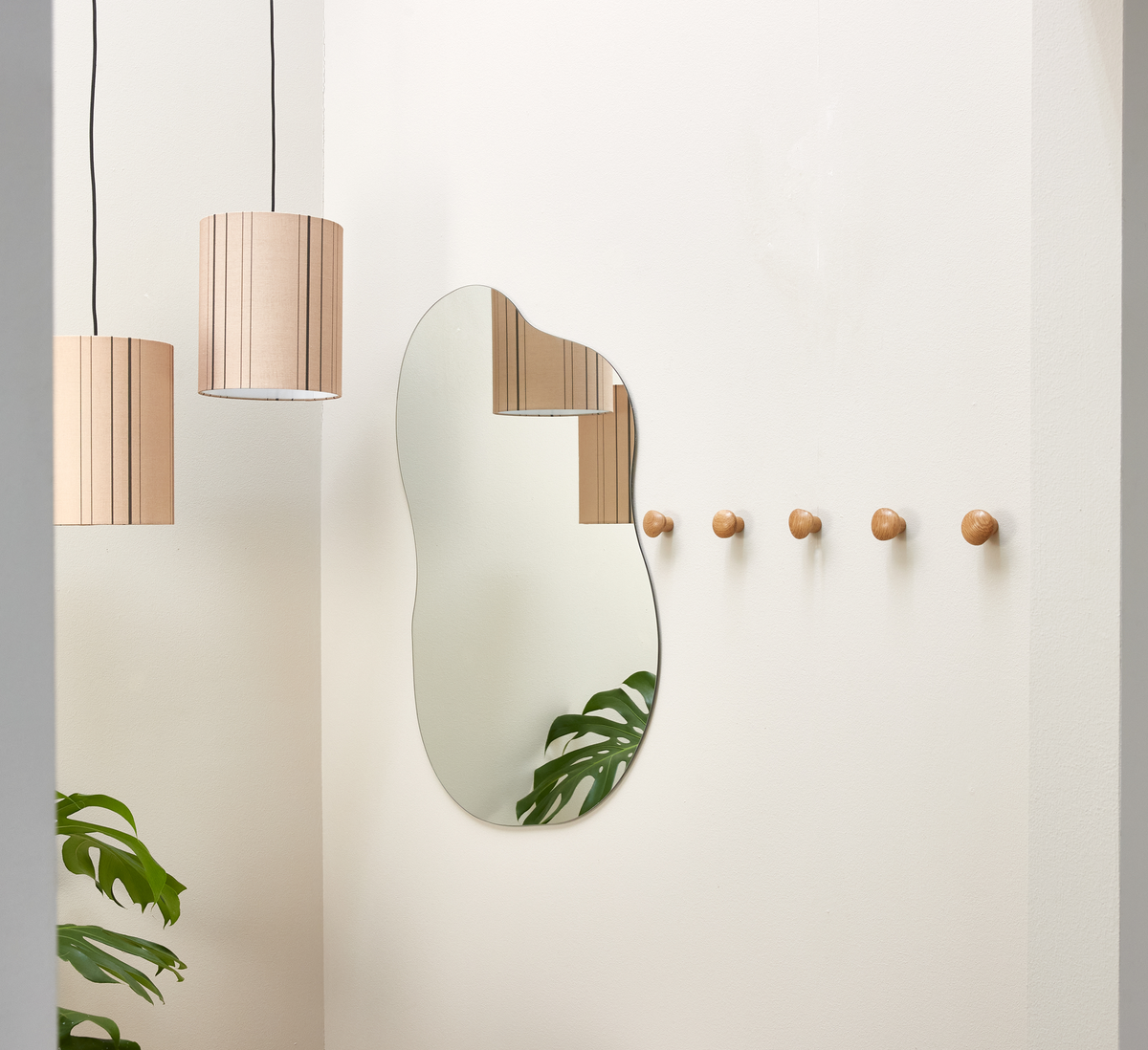 Minimalistisk entré med organisk spejl, række træknopper, to beige pendellamper og monstera-blade mod hvide vægge.