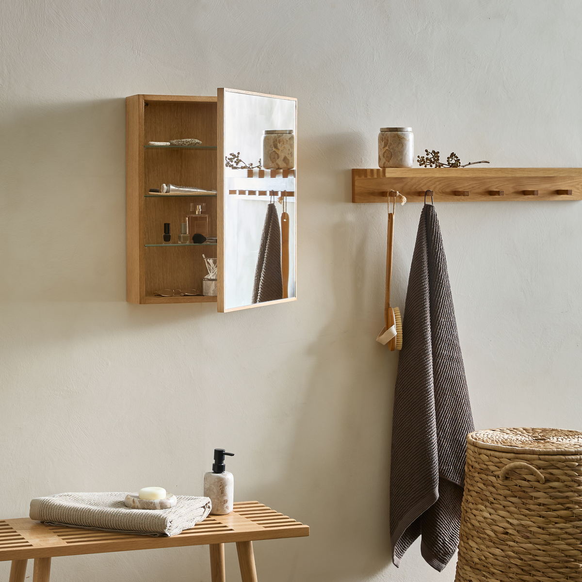 Minimalistisk badeværelsesscene med væghængt spejlskab i træ, knagerække med hængende håndklæde og børste, bænk med sæbe og dispenser samt fletkurv.