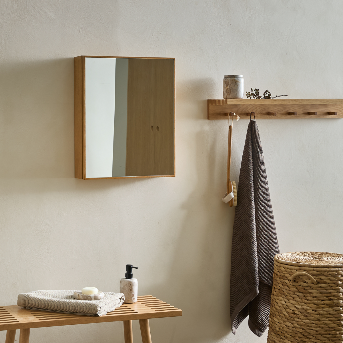 Minimalistisk badeværelsesscene med vægmonteret spejlskab i træ, hylde med knager, hængende mørkt håndklæde og børste, flettet vasketøjskurv samt træbænk med sammenfoldet håndklæde, sæbe og sæbedispenser.